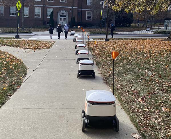Seven food delivery robots in a line travel toward spring street