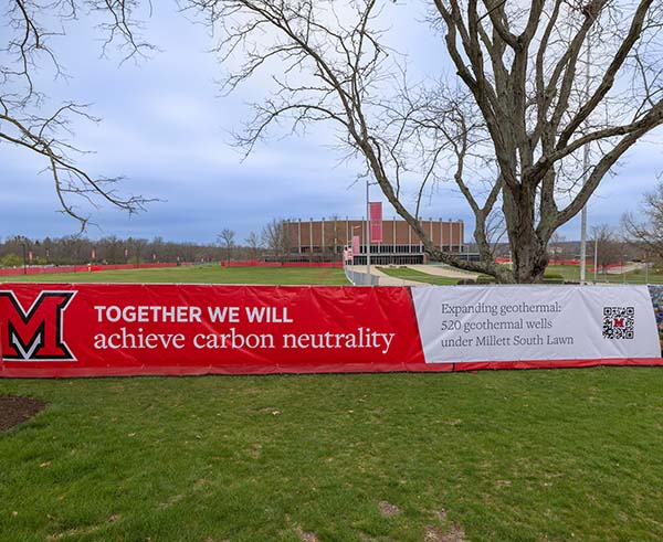 A large banner on a red construction fence  around Millett south lawn with the word Together we will achieve carbon neutrality, and expanding geothermal 520 wells under millet south lawn