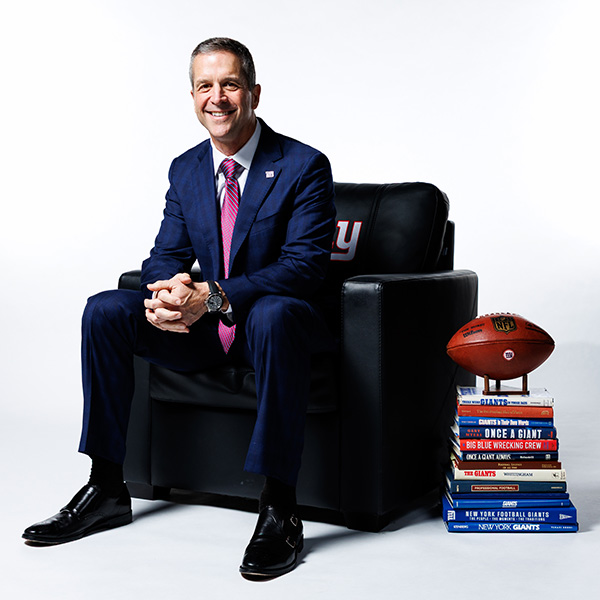 John Harbaugh sitting in a New York Giants branded chair next to a stack of books