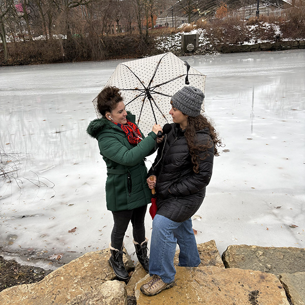 Journalist Rebecca Nagle interviews Kara Strass of the Myaamia Center on the bank of the St. Mary's River in Fort Wayne, Indiana. The river is frozen over and it was a windy day, so they're using an umbrella to block the wind (photo by George Ironstrack).