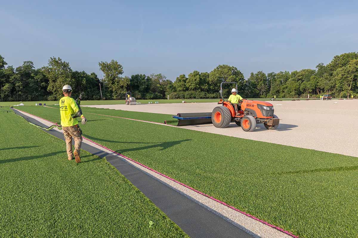 A worker on a tractor unrolls a section of synthetic turf and another worker removes a narrow strip of turf, during the installation of turf at chestnut fields 