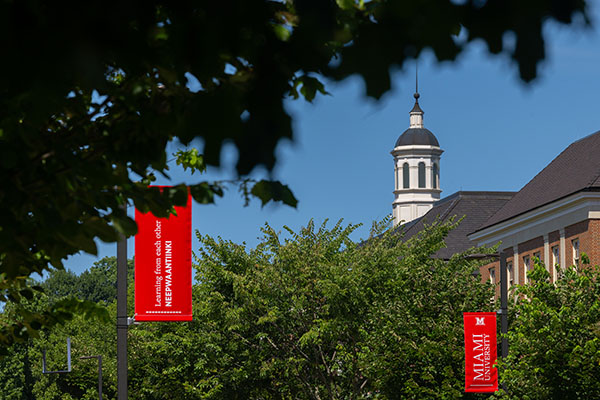 Green leaves of trees frame a blue sky with the Benton Hall cupola in the background and two Miami lightpost banners in the foreground