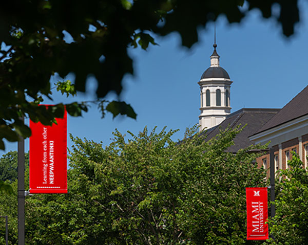Green leaves of trees frame a blue sky with the Benton Hall cupola in the background and two Miami lightpost banners in the foreground