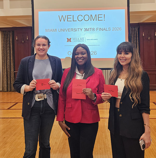 3MT competition winners Faith Dalton, Ruth Ndupu, and Neda Malekikh pose in the Shriver Center Heritage Room each hold a small card indicating their winning rank (second place, first place, second place)