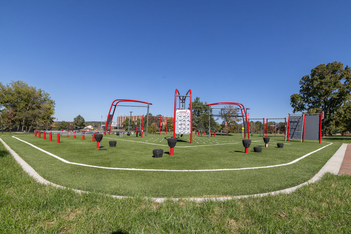 outdoor exercise station with climbing bars, hanging bars, and wooden wall to climb over, with Millett hall in the background