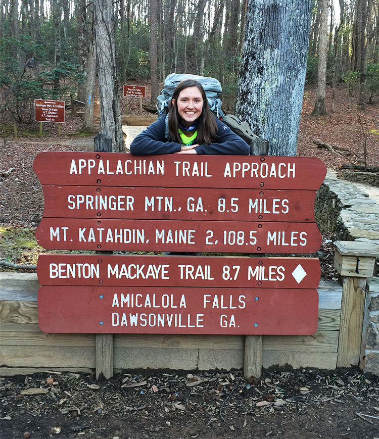 Tess standing behind a sign that says 'Appalachian Trail Approach, Spring Mountain Georgia 8.5 miles, Mt Katahdin, Maine 2108.5 miles'