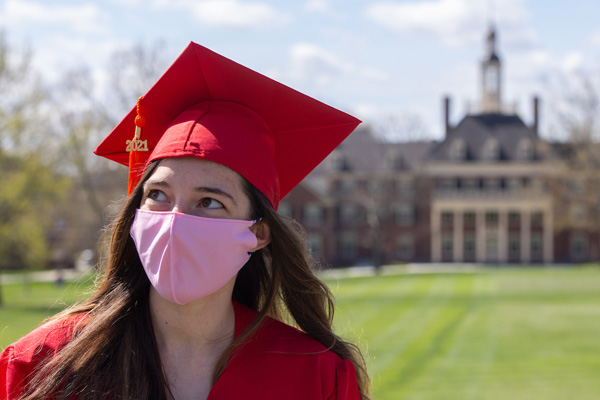 A student in a red graduation cap and gown and pink face mask poses with MacCracken Hall in the background.