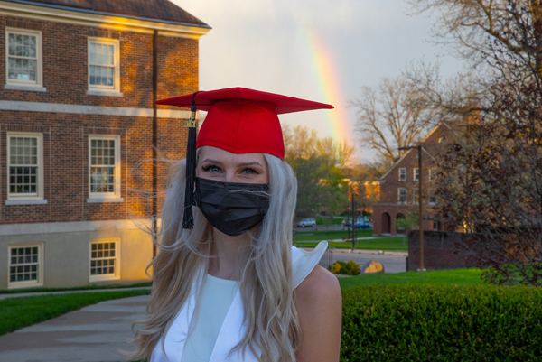 A rainbow and Warfield Hall frame a woman posing with a graduation cap.