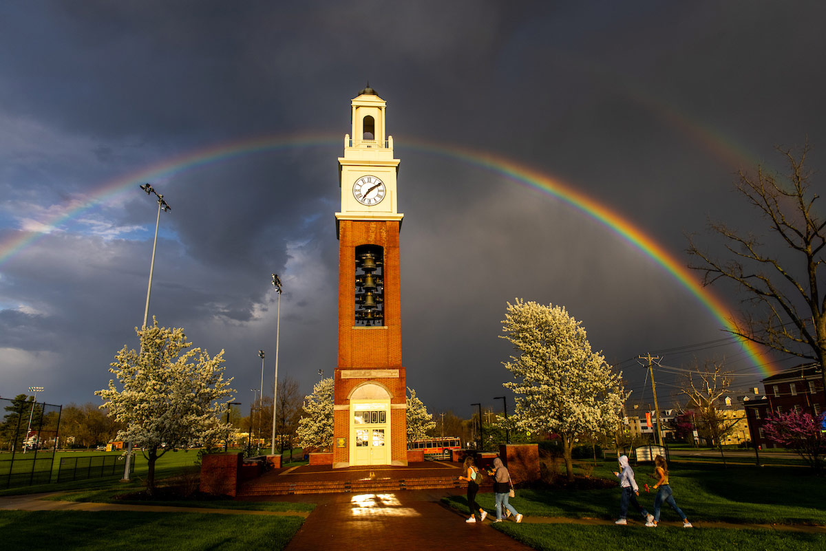 A beautiful rainbow arches over the Pulley Tower, white flowering trees catch the sun rays with dark clouds in the background.