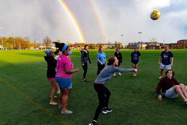A semi-circle of students play a game with a ball on Cook Field with a double rainbow in the background.