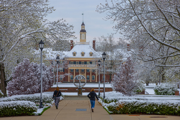 Two students on the path toward the sundial with MacCracken Hall in the background and flowering trees and grass dusted with snow.