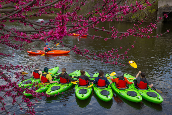 A group of students sit in green kayaks and watch an instructor in a red kayak on Four Mile Creek.