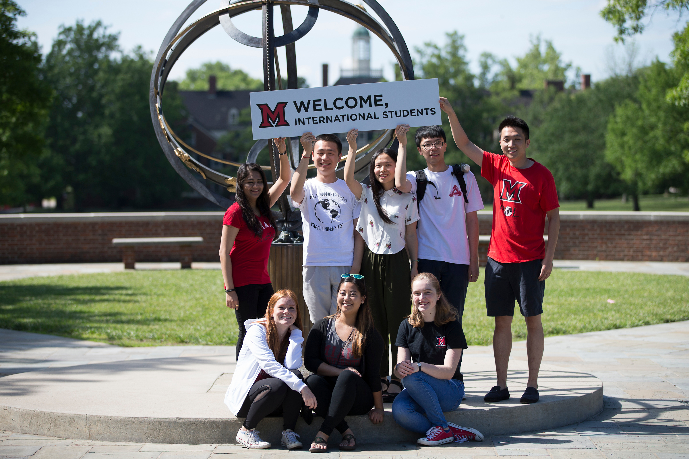 Eight students pose in front of the Tri-Delta Sundial with a sign reading "Welcome International Students."