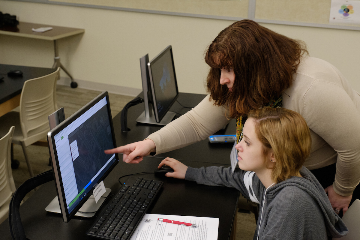 An advisor leans over to show a student something on a computer.