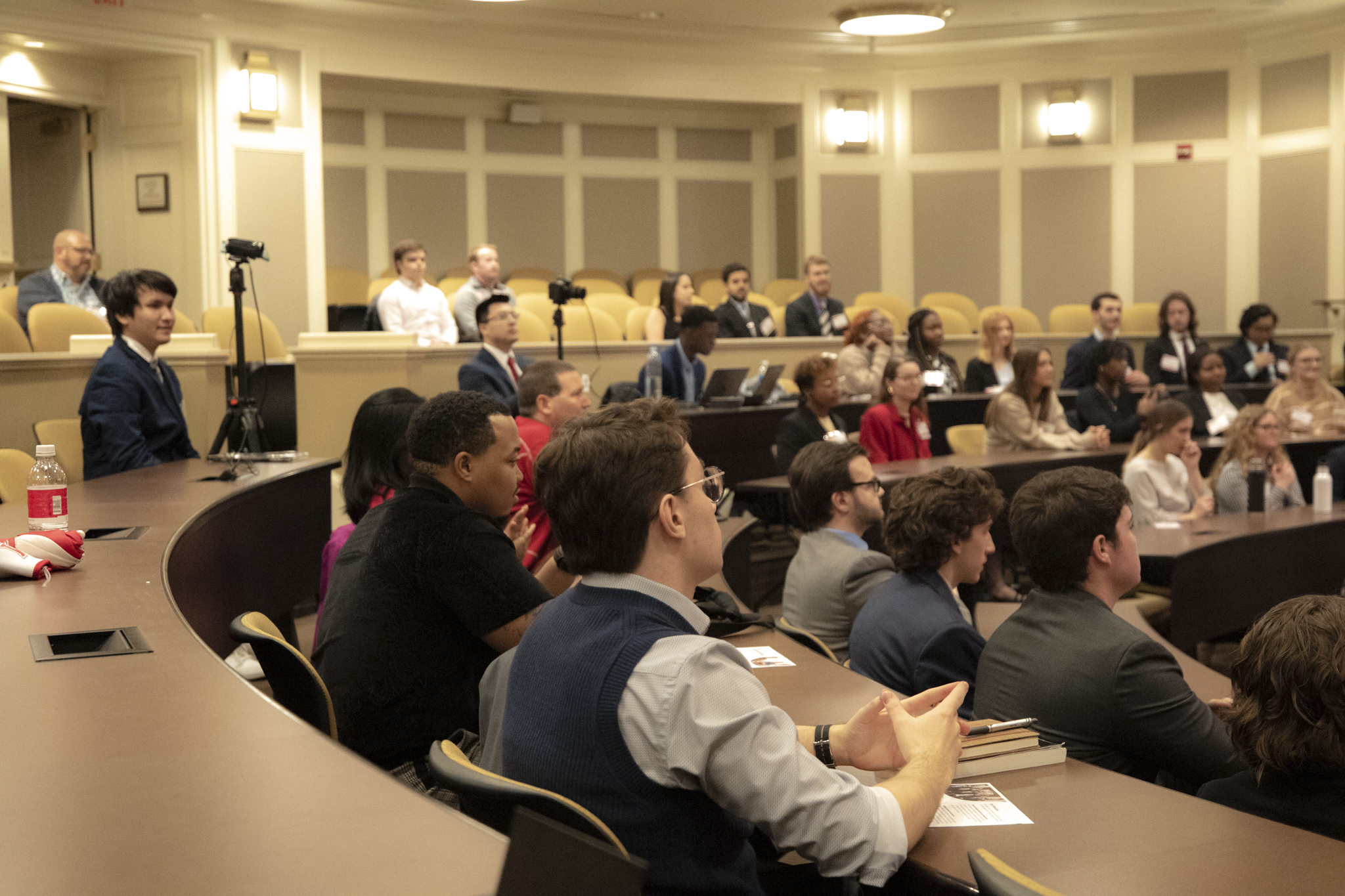 A large group of students sit at tiered tables in a lecture hall.