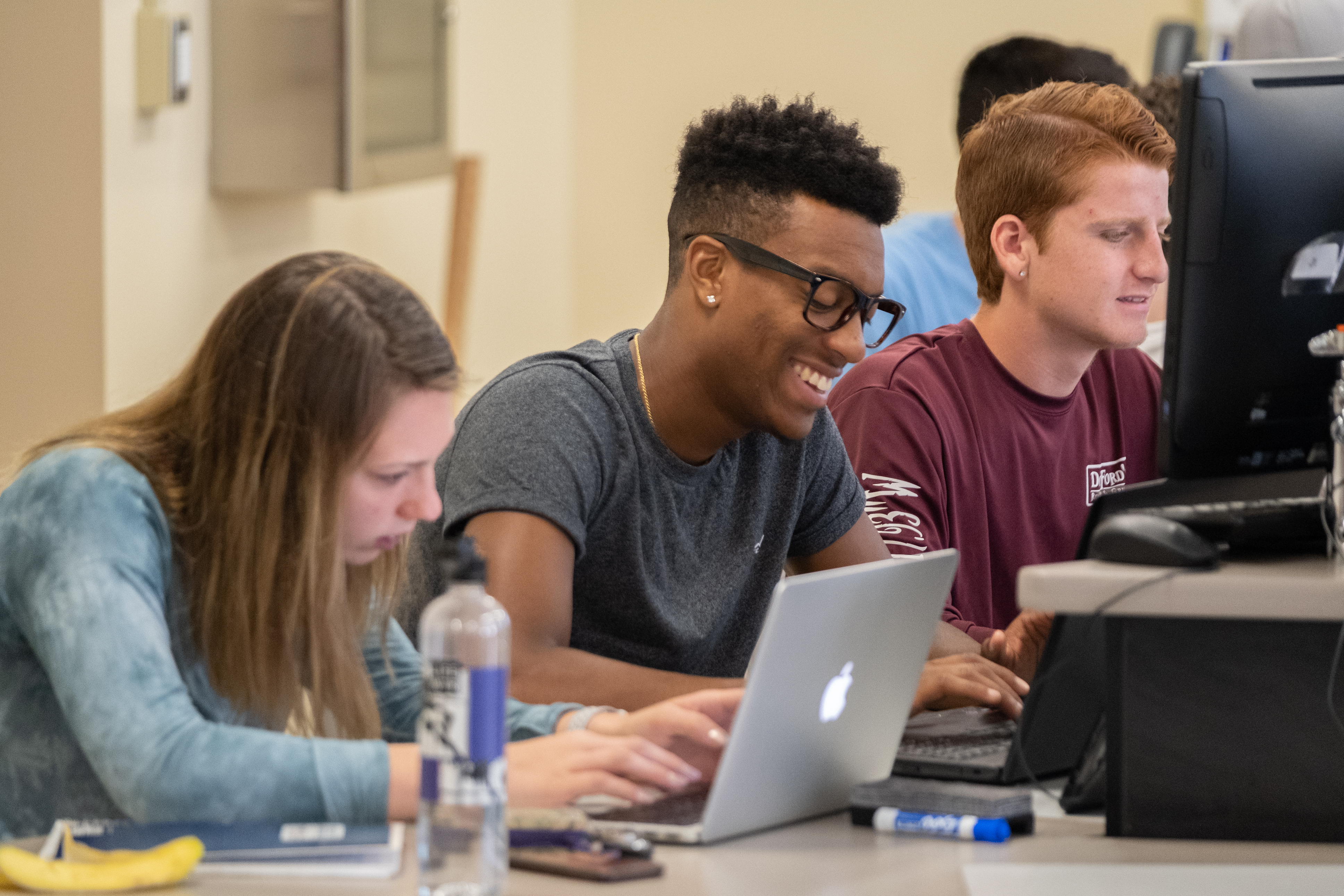 A student smiles while working on a laptop in a computer lab with two peers.