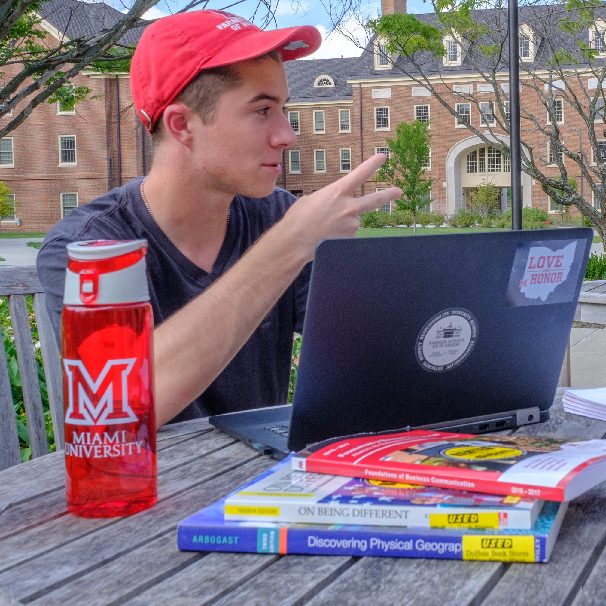Male student with a red Miami hat is sitting outside at a table while working on his computer