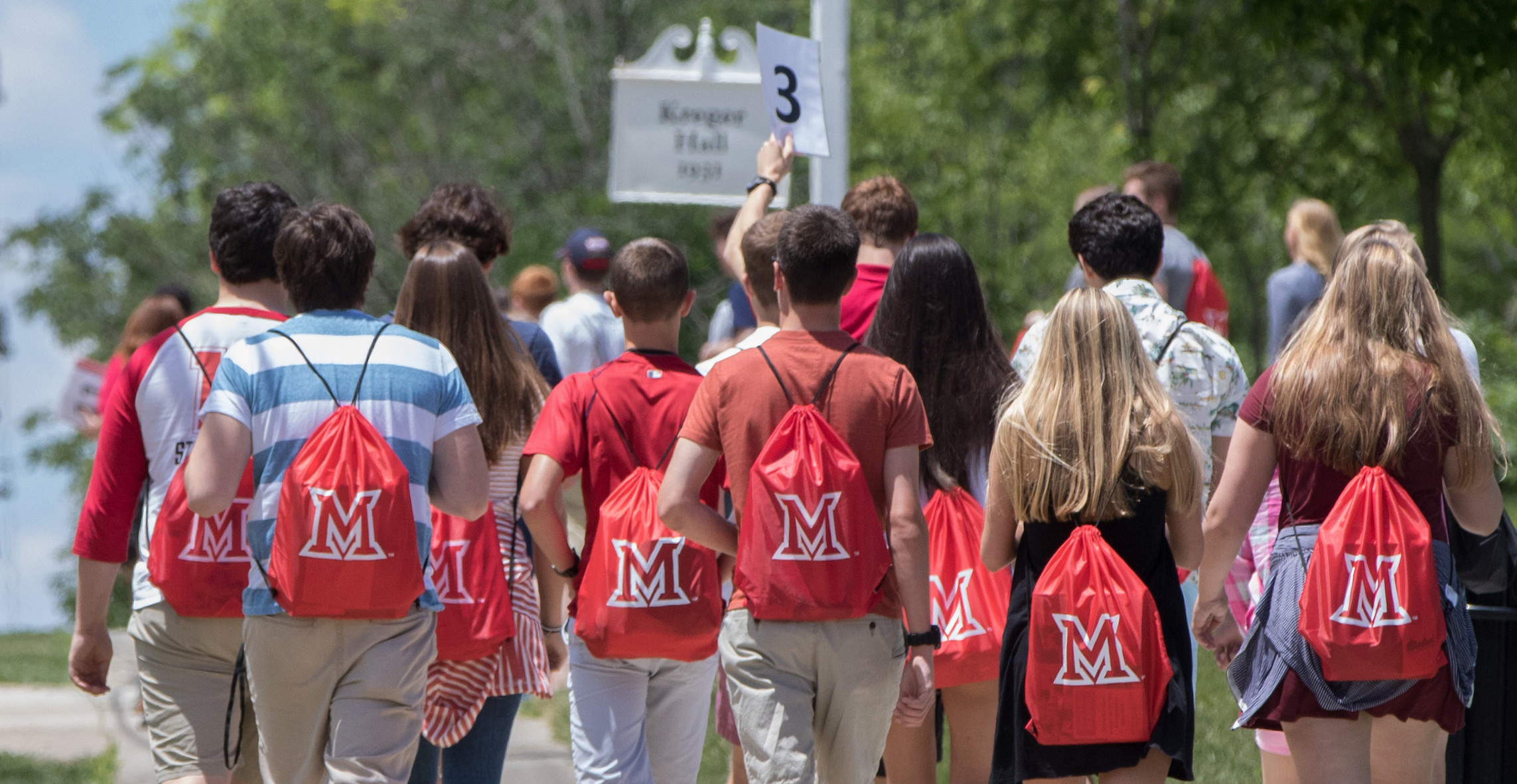 A group of students wearing red Miami drawstring bags walk outside on a sidewalk.