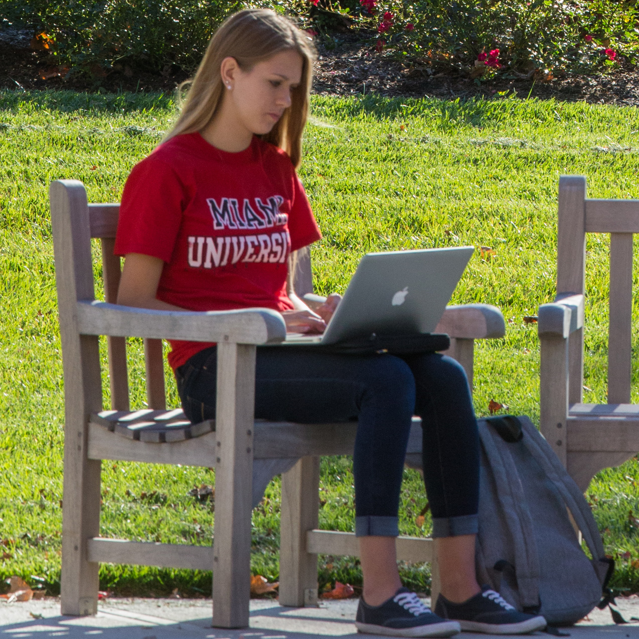 Female student with a red Miami sweatshirt on sitting in a chair outside while working on her laptop.