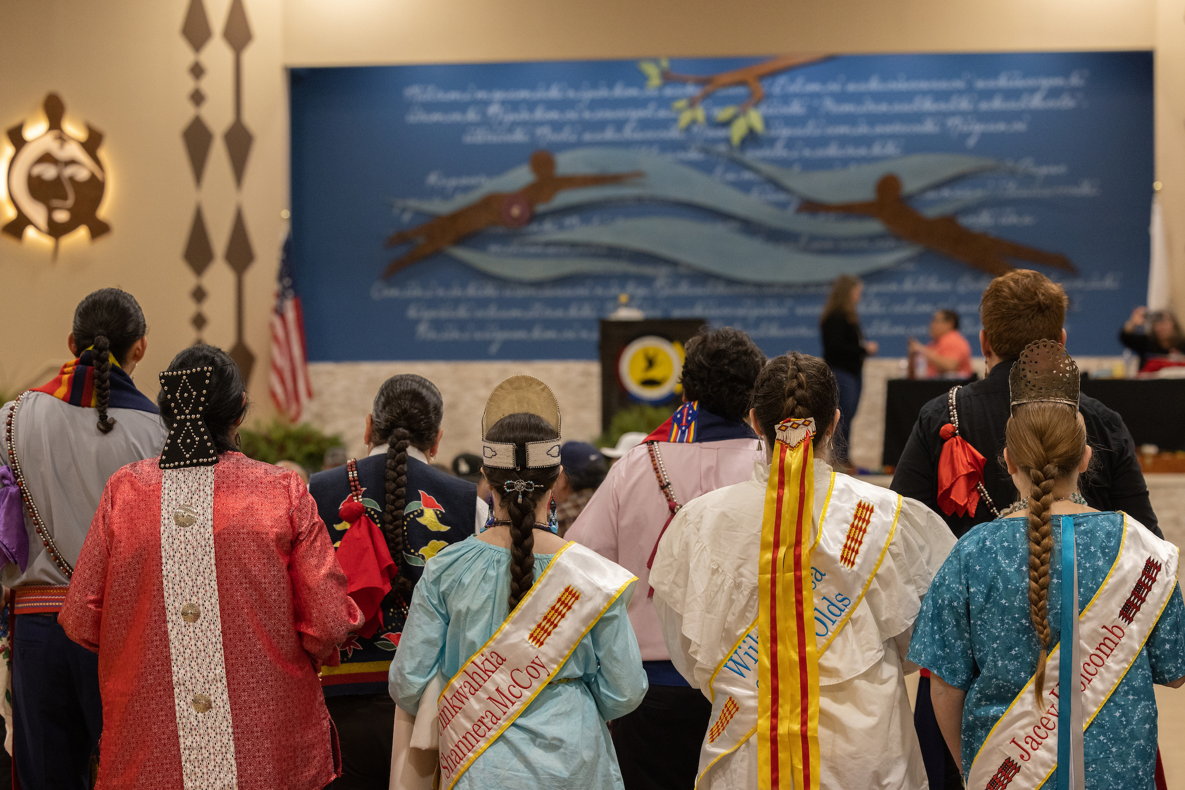 Eight individuals wearing traditional Myaamia attire stand with their backs facing the camera.