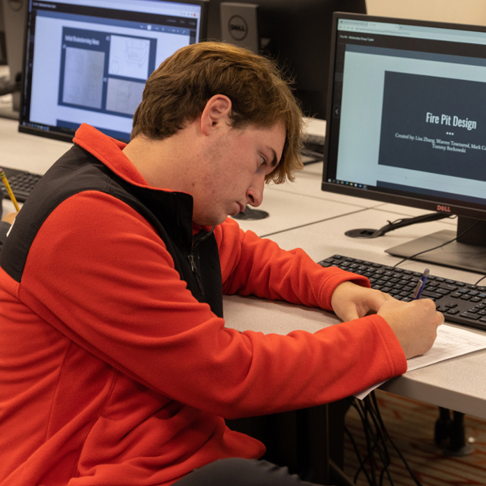 A male student working at a desk with a computer in the background