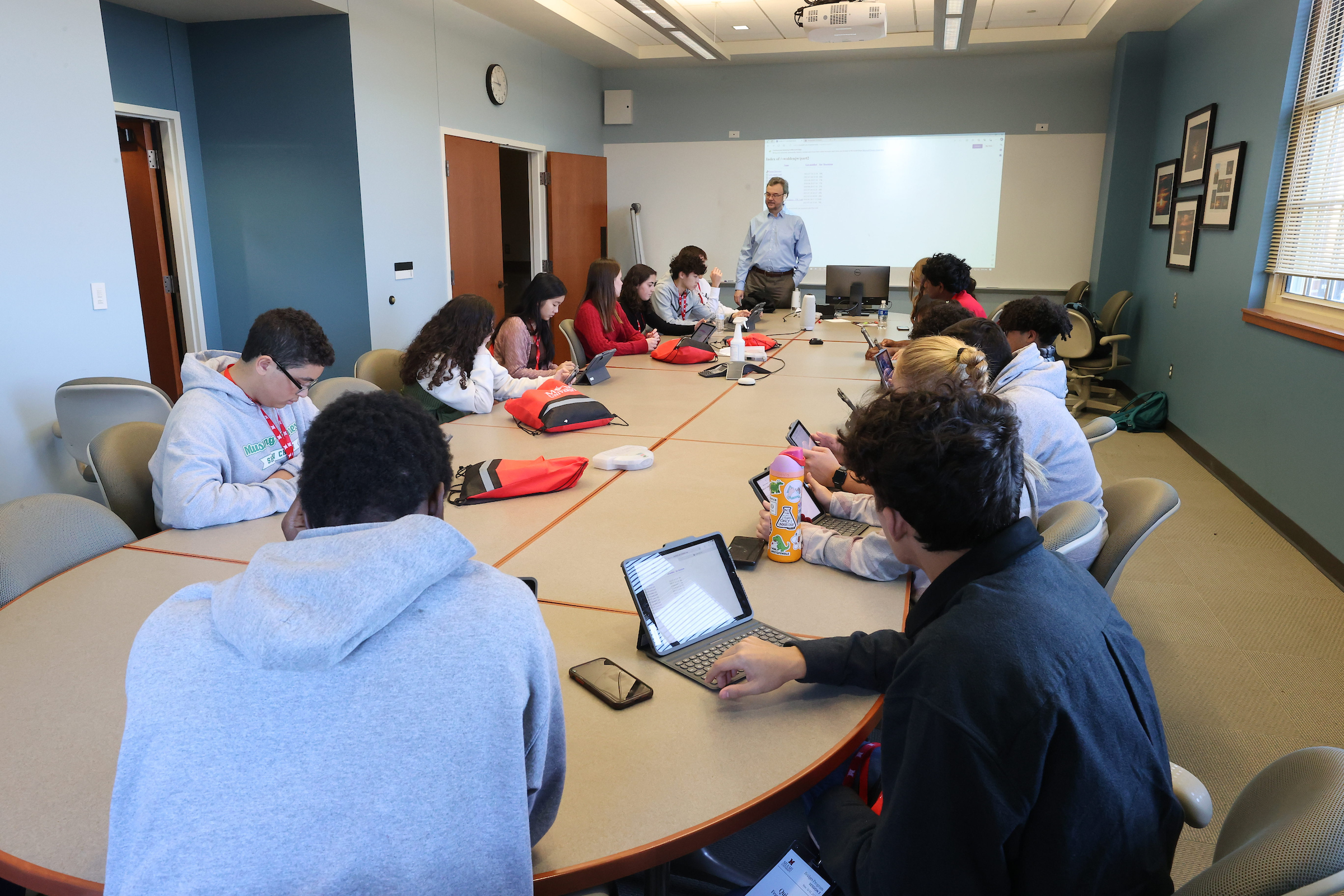 A group of high school students sit at a large table while being addressed by a Miami professor.