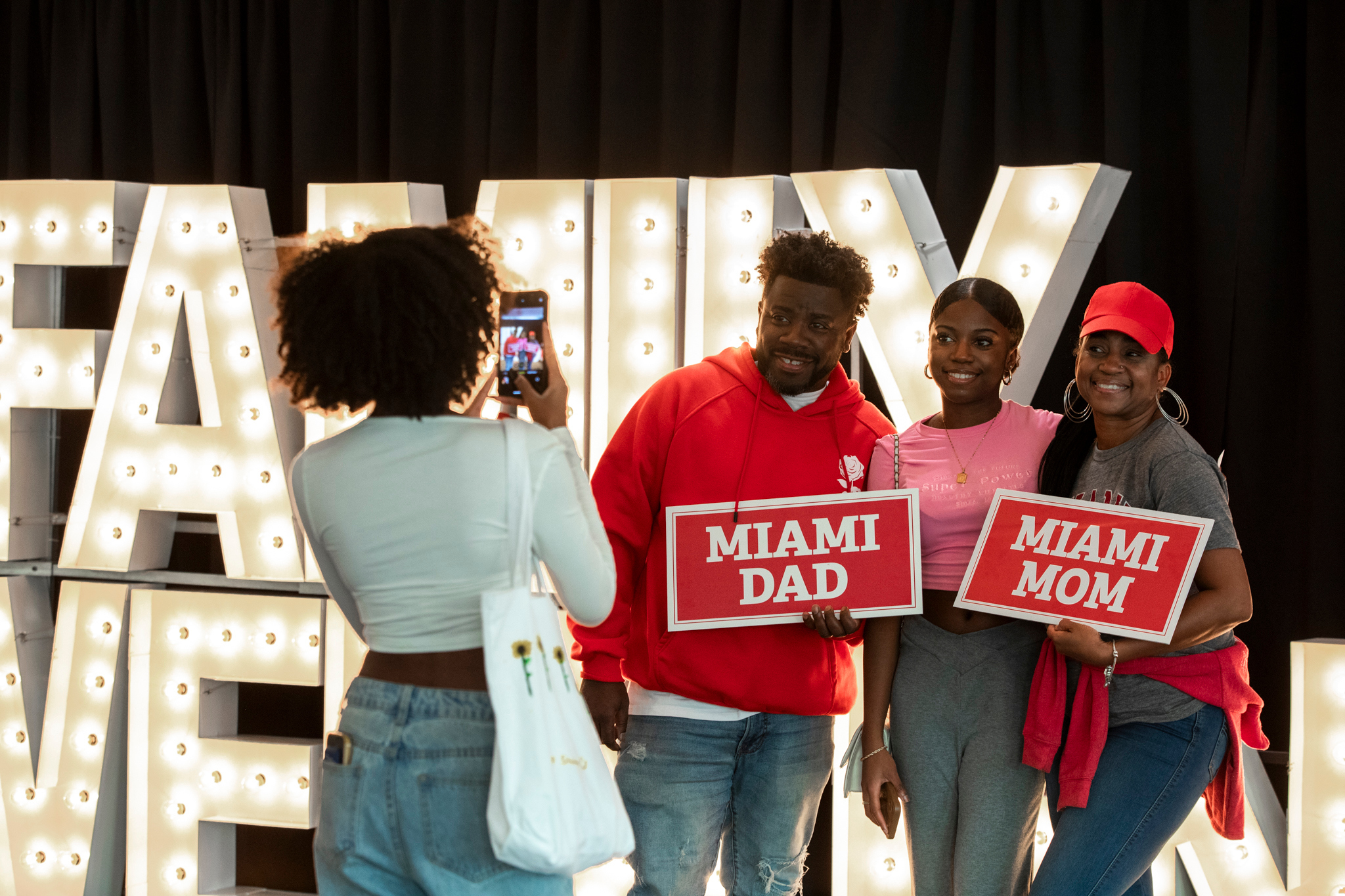 A student and parents pose in front of a "Family Weekend" sign. The parents hold "Miami Mom" and "Miami Dad" signs.