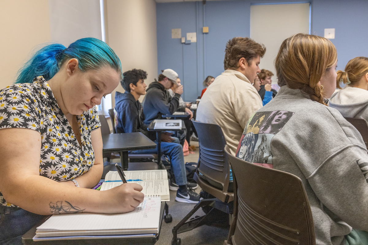 A student takes notes in class while surrounded by peers.