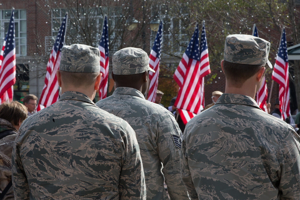 Three soldiers stand with their backs to the camera in front of a row of American flags.
