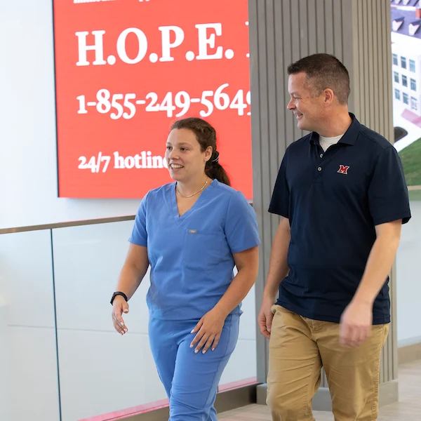 A graduate nursing student walking with her mentor. The H.O.P.E. phone number is listed behind them on a banner.