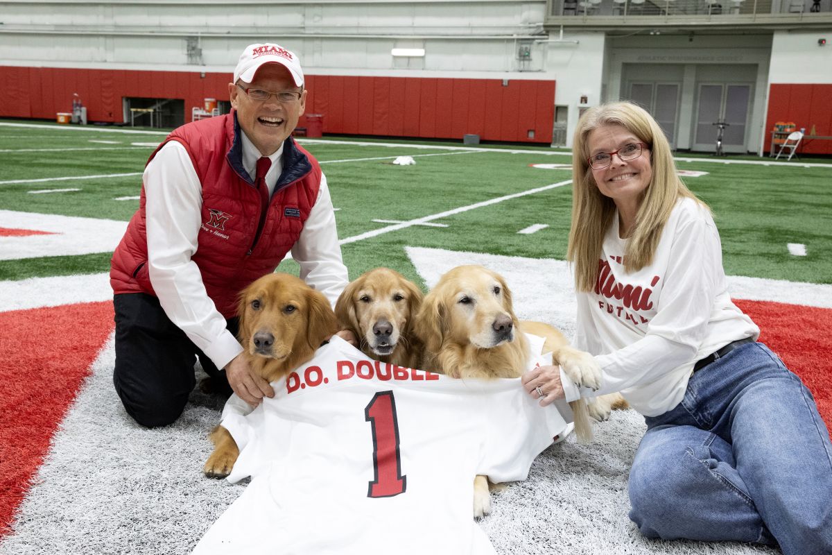 President Greg Crawford and University Ambassador Renate Crawford sit on the Miami University football field with their three dogs while holding a jersey