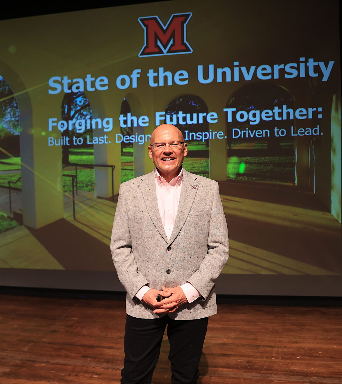 President Crawford stands before his presentation title slide which says State of the University, Forging the Future Together, Built to Last, Designed to Inspire, Driven to Lead.