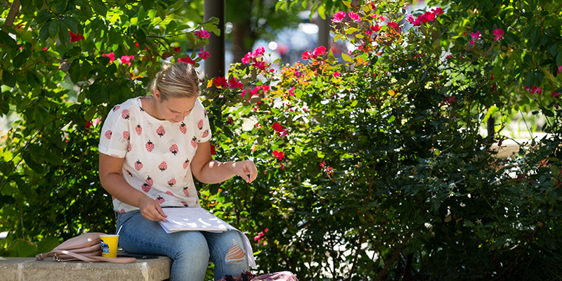 A female student studing in front of pink flowers outide.