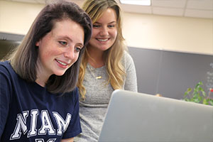 Two females working on a laptop with one sitting down and the other standing looking over her shoulder.