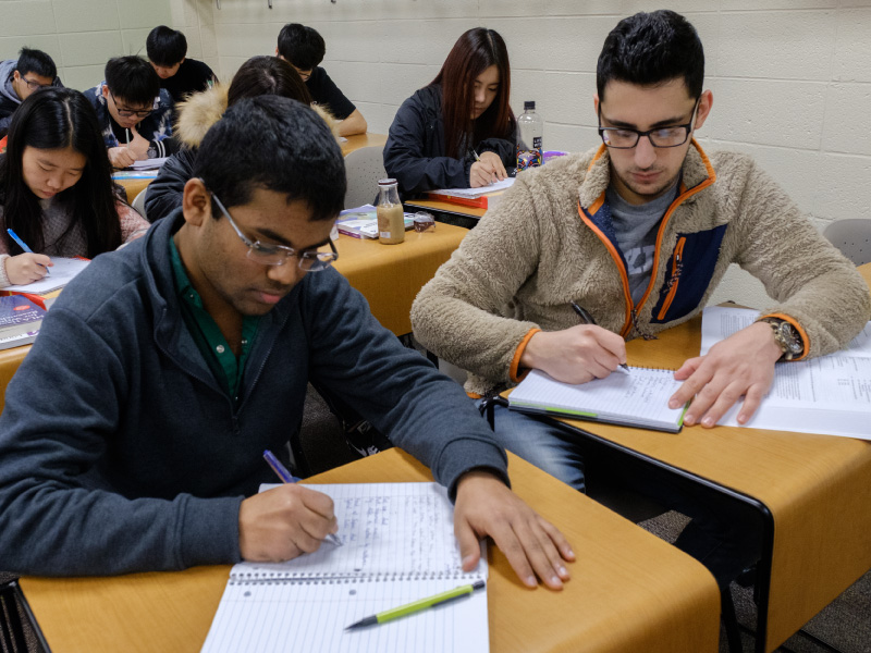 2 students working on at their desks writing on pieces of paper.