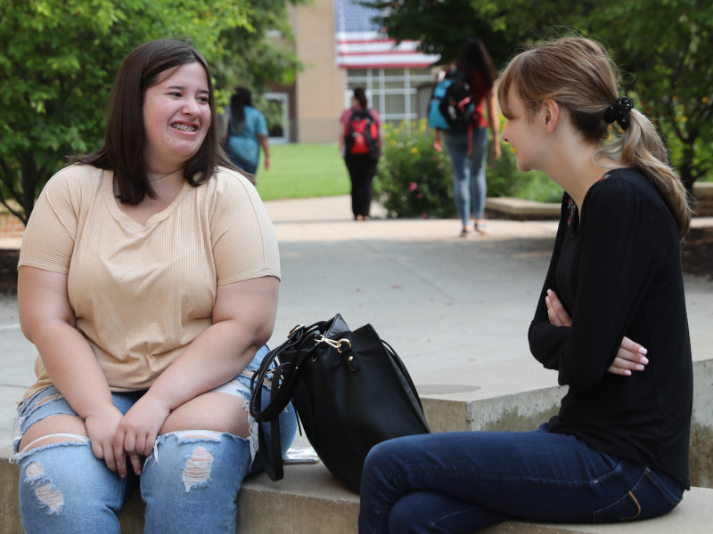 Two students sitting and talking with Miami's campus in the background