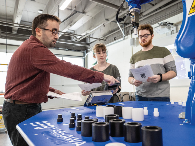 Un profesor les enseña a dos estudiantes a utilizar un robot en el laboratorio de Ingeniería.