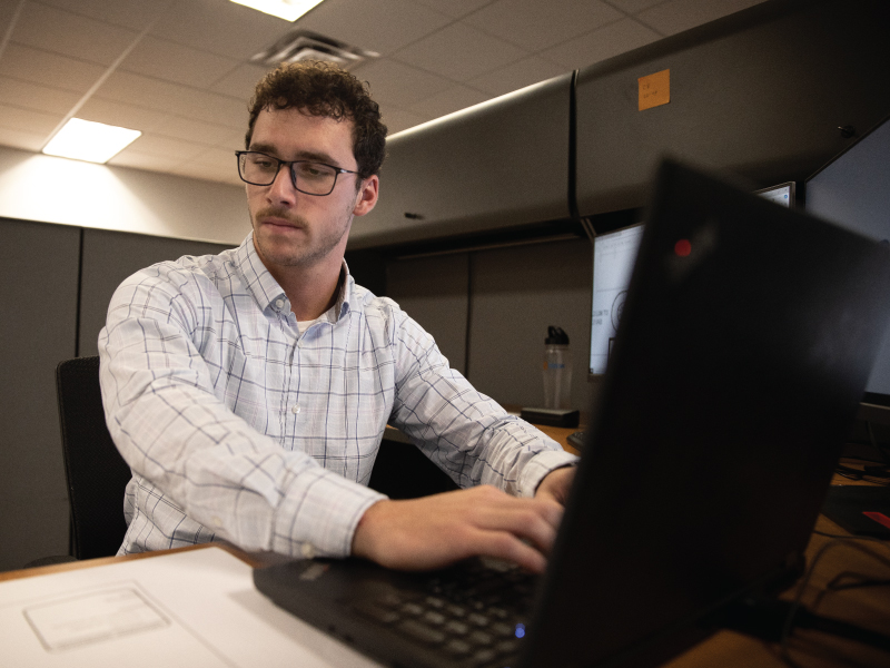 A student looking at a piece of paper on a desk while typing on his computer
