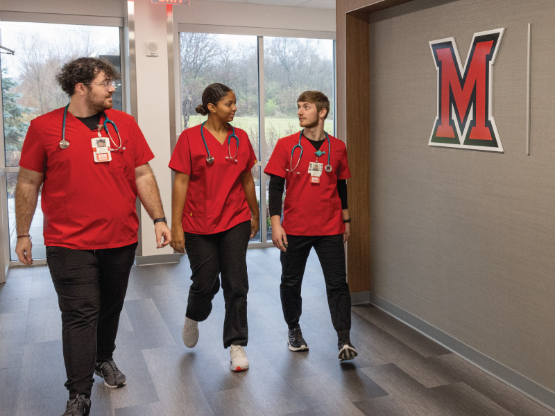 3 Miami nursing students in their scrubs walking down a hallway with a large Miami M on the wall.