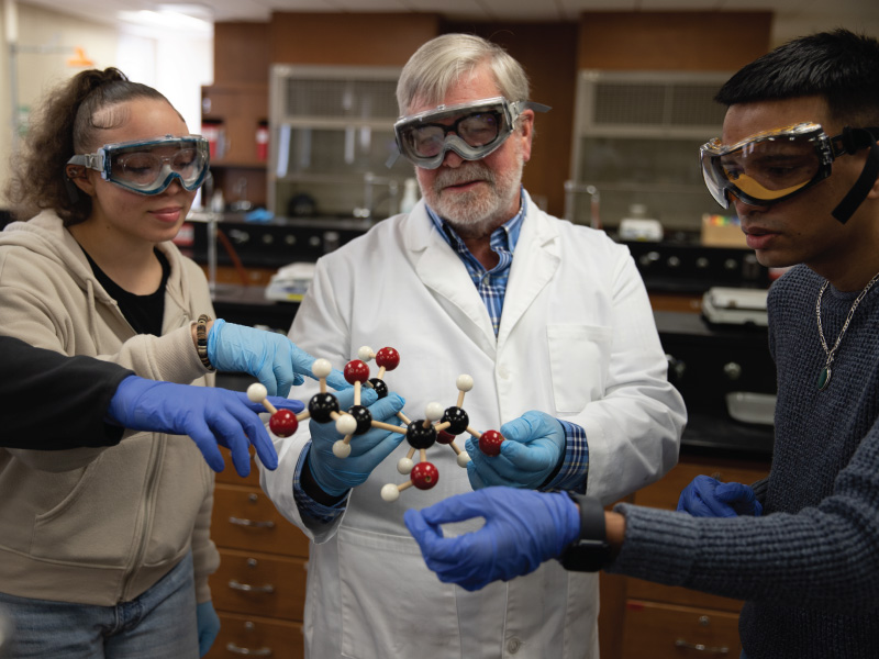 Professor helping 2 students understand a molecular model with a white lab coat and blue gloves.