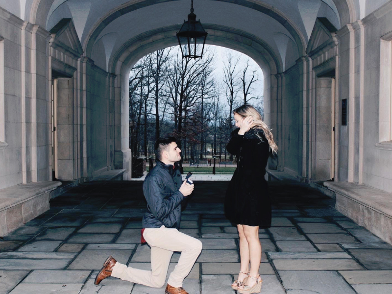 Colin Freistuhler down on one knee holding a ring proposing to to Delaney O'Brien under the Upham Hall Arch.