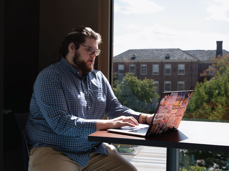 Zachary Matthews working on his laptop sitting by a window with Oxford buildings in the background. 
