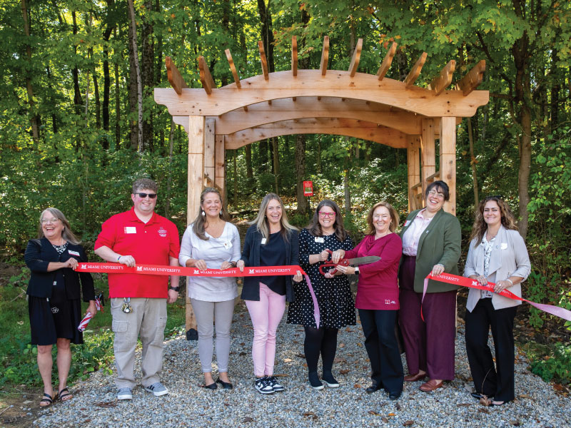 Individuals holding a ribbon with Moira Casey holding scissors infront of the entrance of the Haiku trail.