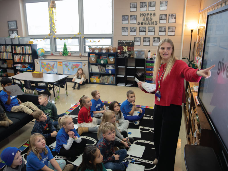 Jessica Goodwin standing in front of the class who are sitting on a carpet pointing at the smart board.