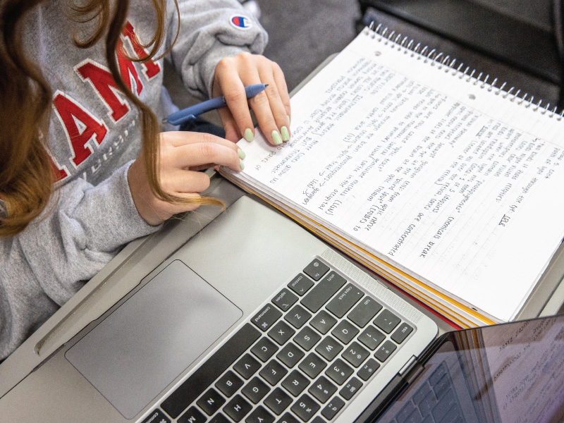 Student taking notes in her notebook with her laptop on her desk.