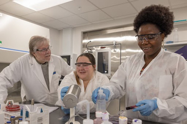 Three laboratory technicians in white lab coats and safety goggles collaborate on an experiment, pouring substances into a container while discussing their work.