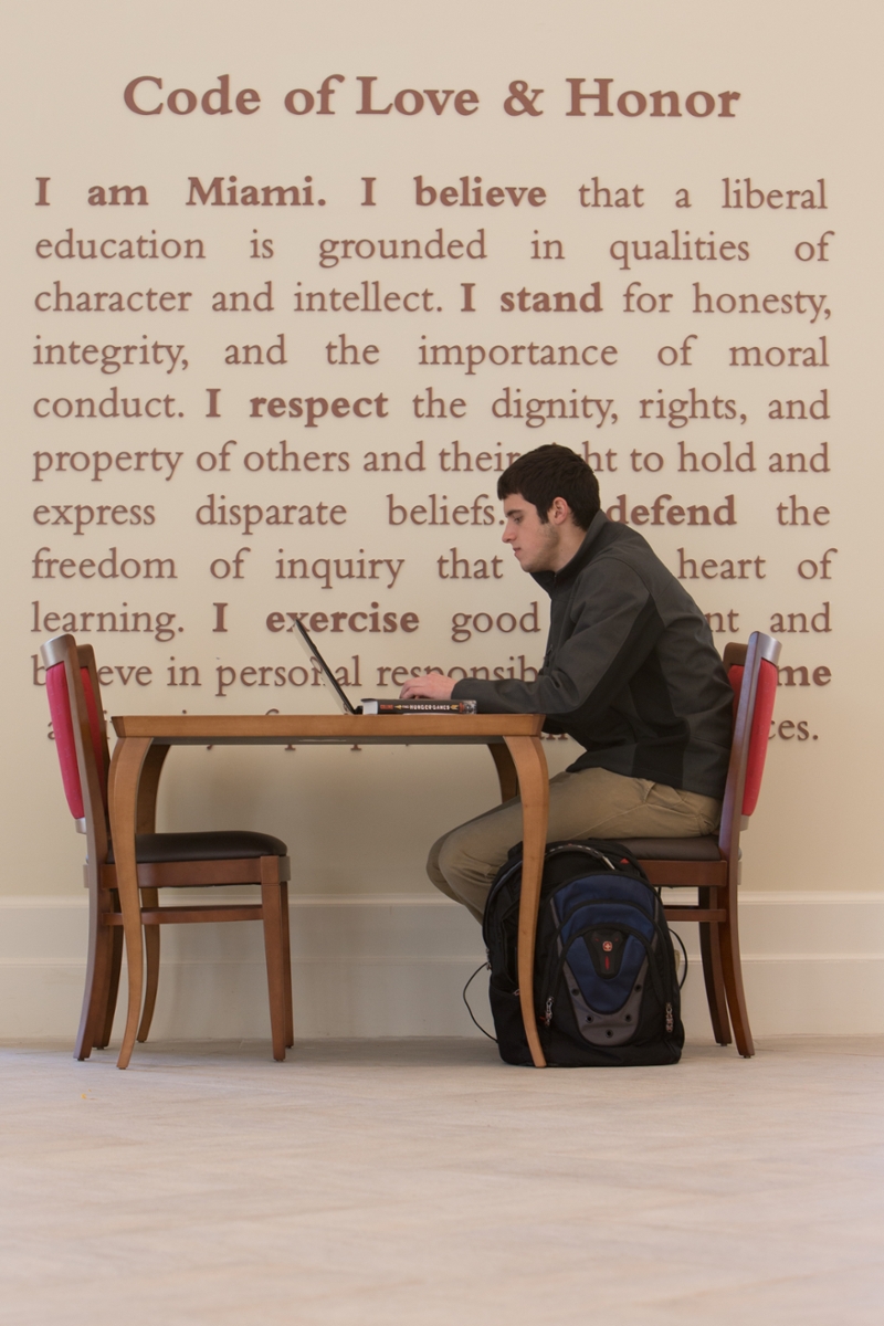 Wall in Armstrong that reads the Code of Love and Honor, with a male student sitting at a desk and looking at laptop in front of it. 