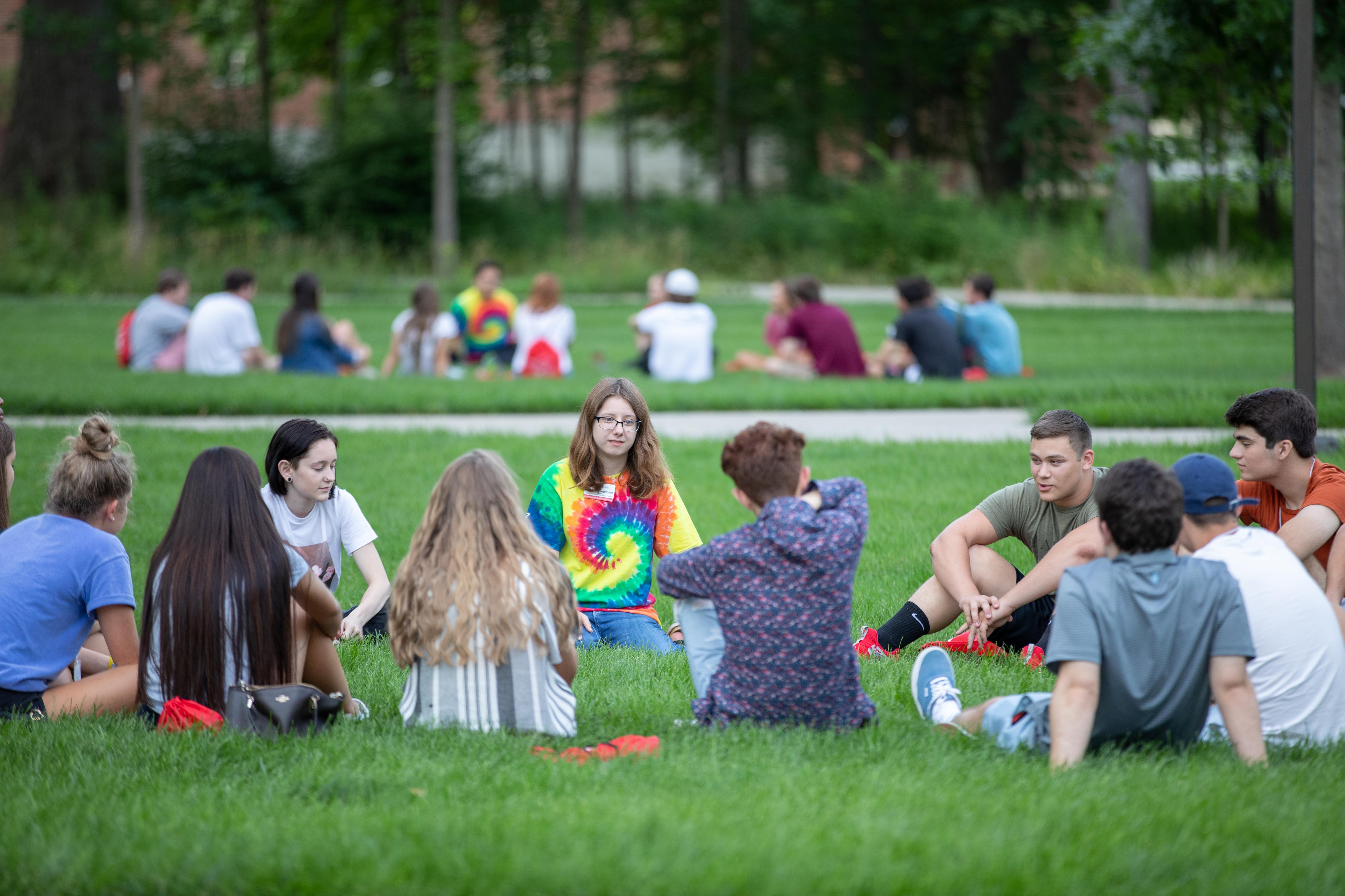 Two groups of about 15 students sitting in circles on a grassy area of campus for orientation.