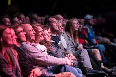 An audience in a theater looking up and laughing at a performer off camera