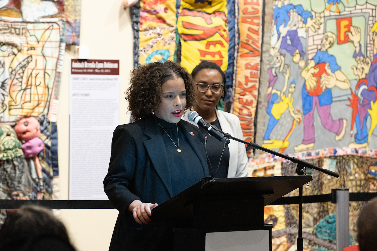 Nyah Smith stands at a podium speaking into a microphone, while Jules Jefferson stands behind her. 